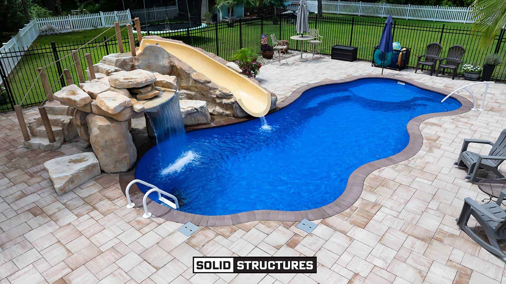 RicoRock, GROTTO AND WATER SLIDE: SR Smith BigRide Water Slide Right Turn 5.5 Feet-Taupe-BR-2L-TP with PKG 15 from, PAVER WALKWAY: Belgard 2'x3' Slabs COLOR: Sandbridge, COPING: Belgard Marina Coping COLOR: Chocolate, FENCE:6' Tall Black Aluminum Fence with Through Pickets, ROCKBED: 1-3" Brown/White/Tan River Rock with Landscaping Fabric Beneath, 6' Tall White Vinyl Privacy Fence with A 10' Wide Gate (Two 5' Gate Leaves Each) and a 5' Wide Gate, PAVERS: Belgard Dimensions 12 COLOR: Sandbridge Standard Finish with Chocolate 6x9 Accent Stones in Soldier Course, POOL: Thursday Pools 36' Wellspring COLOR: Maya