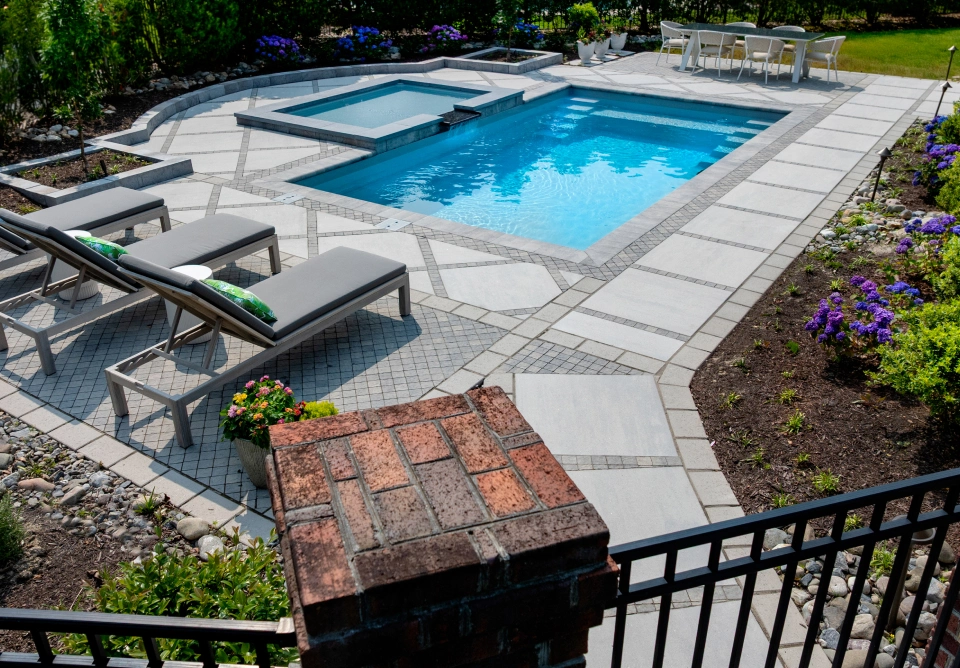 Backyard pool with an attached spa, gray stone patio, and two sun loungers nearby with a dining table in the distance.