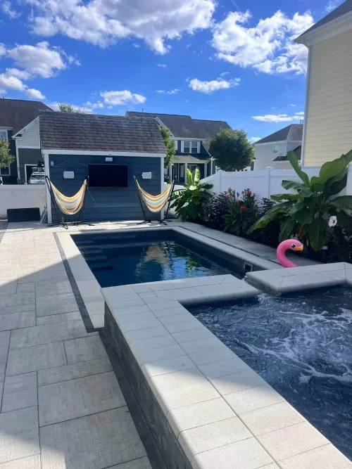 3 after Compact Pool and Spa Construction - Solid Structures Backyard Oasis with a Rectangular Pool and Raised Spa, Pink Flamingo Float, Two Striped Hammocks on a Gray Shed, and Lush Plants Along a White Fence on a Sunny Day.