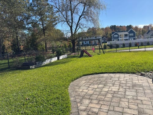 5 Before Backyard Transformation with Pool and Outdoor Living in Great Bridge - Solid Structures Sunlit Park Lawn with a Curved Stone Patio, a Small Playground with a Slide Beyond a Black Fence, and Houses in the Background.