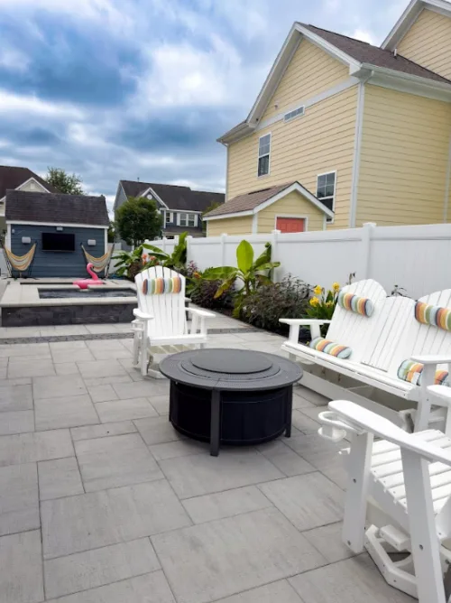 6 after Compact Pool and Spa Construction - Solid Structures Patio with White Adirondack Chairs and Striped Cushions Around a Round Black Fire Pit on a Gray Tile Deck, Yellow House in Background.