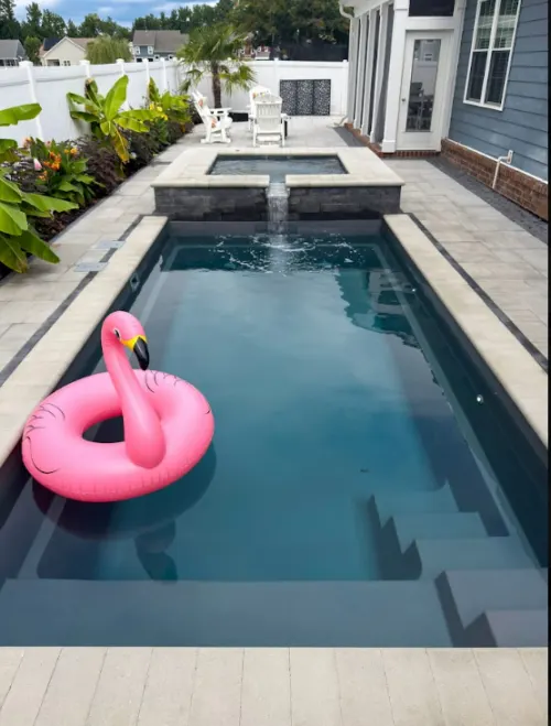 7 after Compact Pool and Spa Construction - Solid Structures Rectangular Backyard Pool with a Spa at the Far End, Pink Flamingo Float on the Water, and Lounge Chairs by a White Fence.