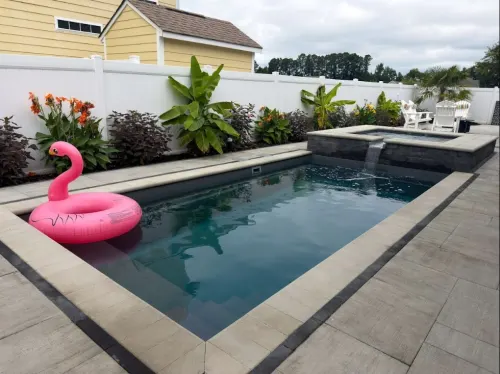 After Compact Pool and Spa Construction - Solid Structures Backyard Rectangular Pool with a Pink Flamingo Float, Surrounded by a Concrete Deck and White Fence, Tropical Plants Along the Edge.