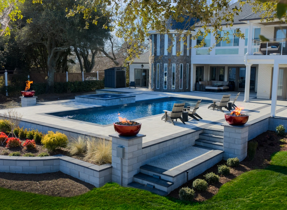 Elegant backyard with a rectangular pool, spa, and stone patio; fire bowls glow on pedestals by the steps.