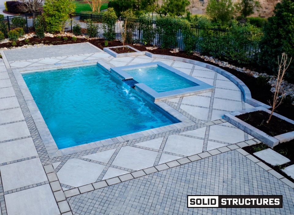 Elegant backyard with a rectangular pool, spa, and stone patio; fire bowls glow on pedestals by the steps.