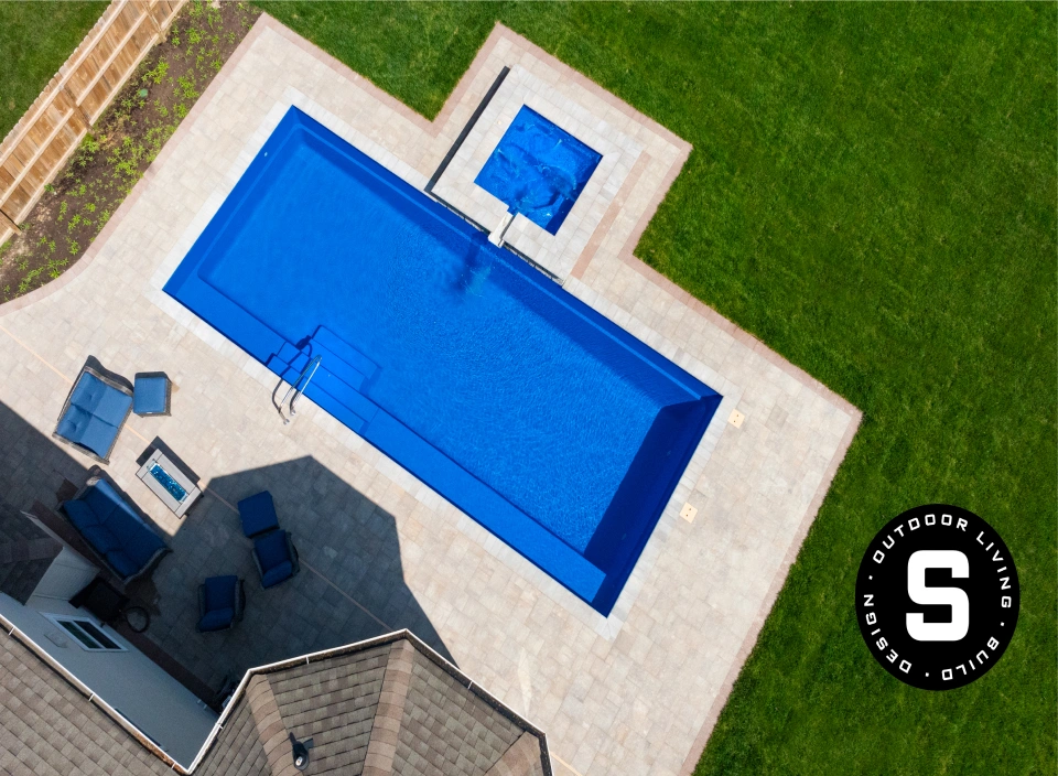 Aerial view of a rectangular blue swimming pool on a tiled patio, with a square spa beside it, blue lounge chairs, and surrounding green lawn; logo in the bottom-right corner.