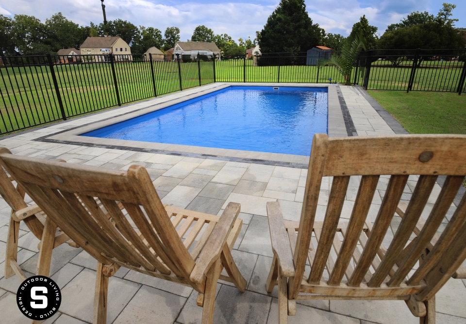 Rectangular blue pool bordered by light gray paver patio and a black metal fence, with two wooden lounge chairs in the foreground.