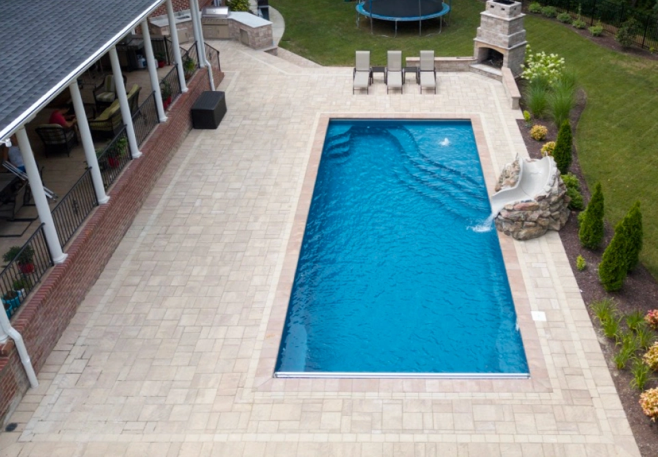 A rectangular blue swimming pool with a beige tiled pool deck, adjacent patio seating under a covered porch on the left, and landscaped yard with a trampoline in the background.