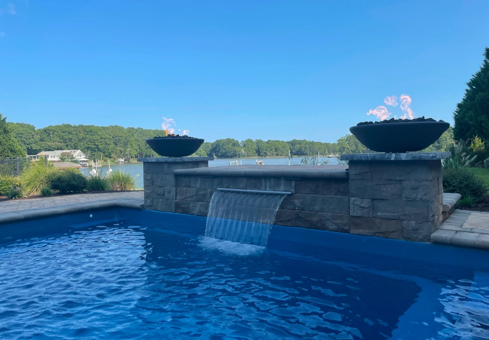 Backyard pool with a stone waterfall feature and two fire bowls on pedestals overlooking a lake and trees.