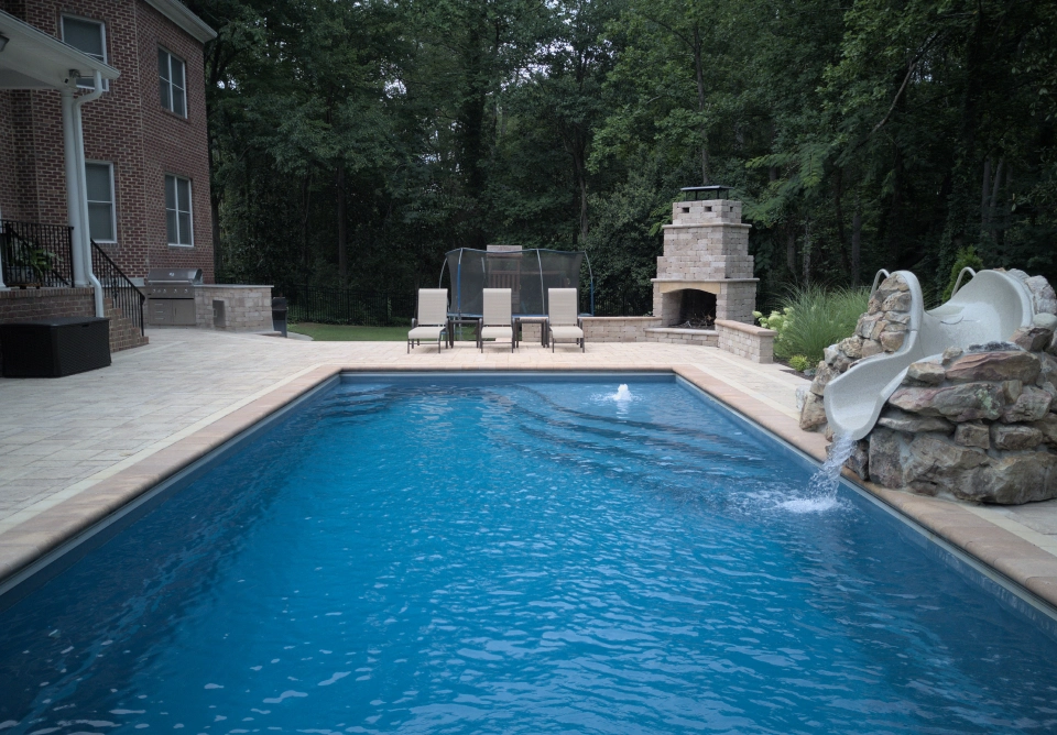Backyard pool area with a rectangular blue pool, stone deck, and a right-side rock waterfall slide near a built-in fireplace.