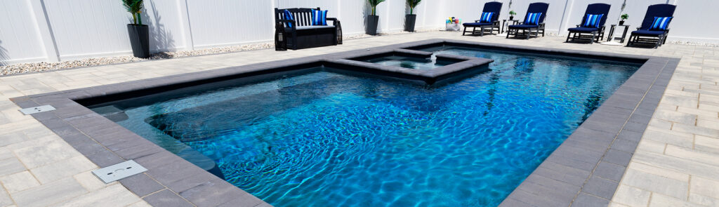 Rectangular In-ground Pool with a Raised Spa Feature, Surrounded by a Light Stone Patio and Blue-cushioned Lounge Chairs.