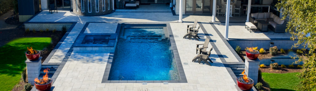 a Modern Backyard Featuring a Rectangular Pool with a Connected Spa, Two Red Fire Bowls on Pedestals at the Sides, and a Gray Stone Patio with Lounge Chairs and Seating Area.