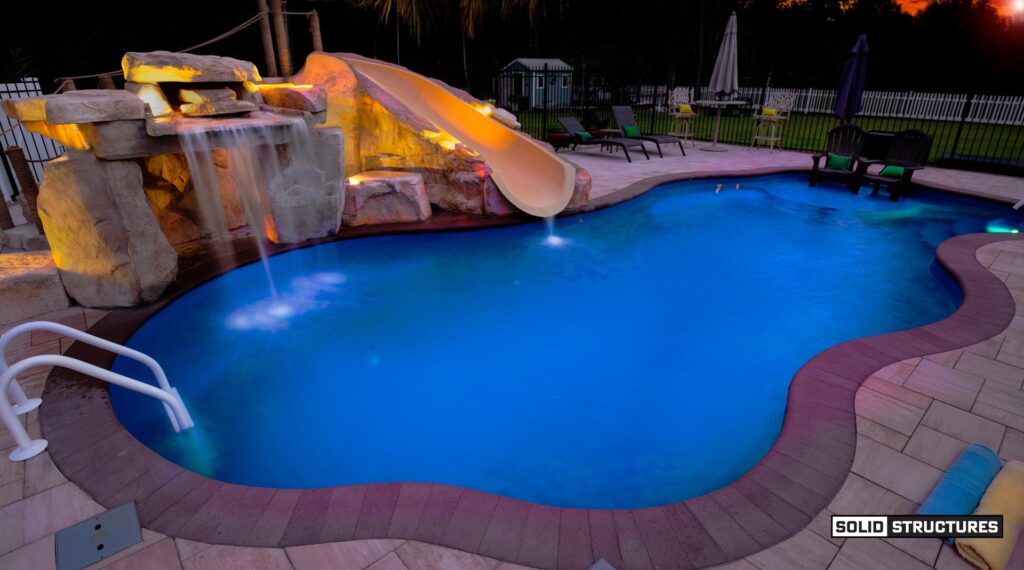 Ricorock, Grotto and Water Slide: Sr Smith Bigride Water Slide Right Turn 5.5 Feet-taupe-br-2l-tp with Pkg 15 From, Paver Walkway: Belgard 2'x3' Slabs Color: Sandbridge, Coping: Belgard Marina Coping Color: Chocolate, Fence:6' Tall Black Aluminum Fence with Through Pickets, Rockbed: 1-3" Brown/white/tan River Rock with Landscaping Fabric Beneath, 6' Tall White Vinyl Privacy Fence with a 10' Wide Gate (two 5' Gate Leaves Each) and a 5' Wide Gate, Pavers: Belgard Dimensions 12 Color: Sandbridge Standard Finish with Chocolate 6x9 Accent Stones in Soldier Course, Pool: Thursday Pools 36' Wellspring Color: Maya